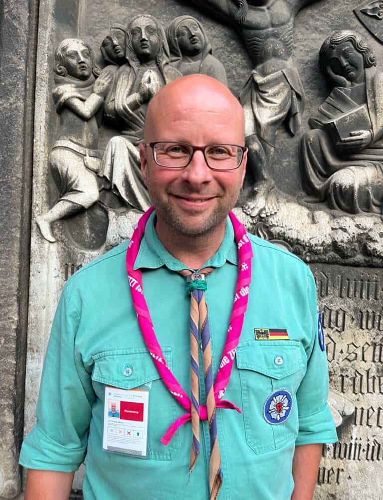 Ein Mann mit Brille und pinkem Halstuch vor einer historischen Steinmetzarbeit, lächelnd.