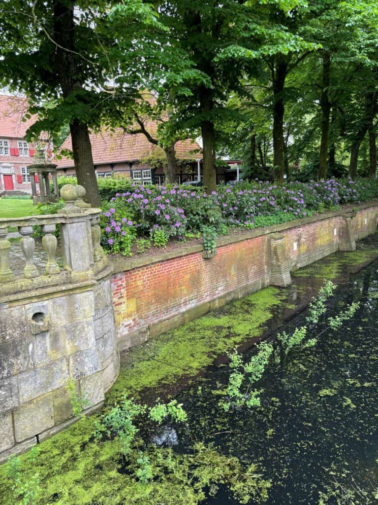 Grüner Park mit Wasser, Bäume und Blumen, historische Stadtmauer im Hintergrund.