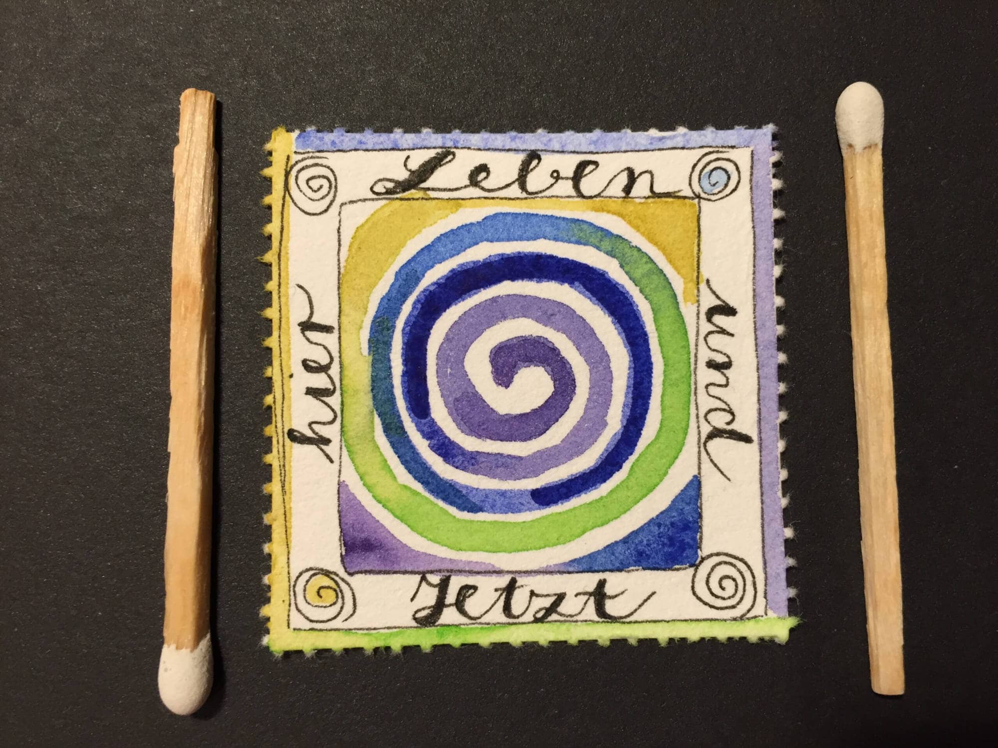 Buntes spiralförmiges Kunstwerk auf Stoff mit Holzstäbchen an den Seiten.