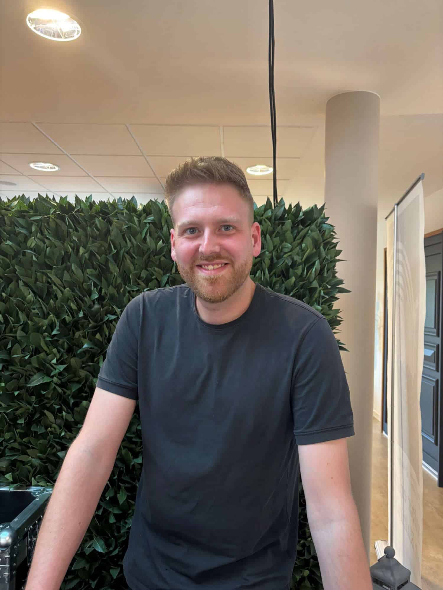 Freundlicher junger Mann mit Bart vor grüner Pflanzenwand in Innenraum.